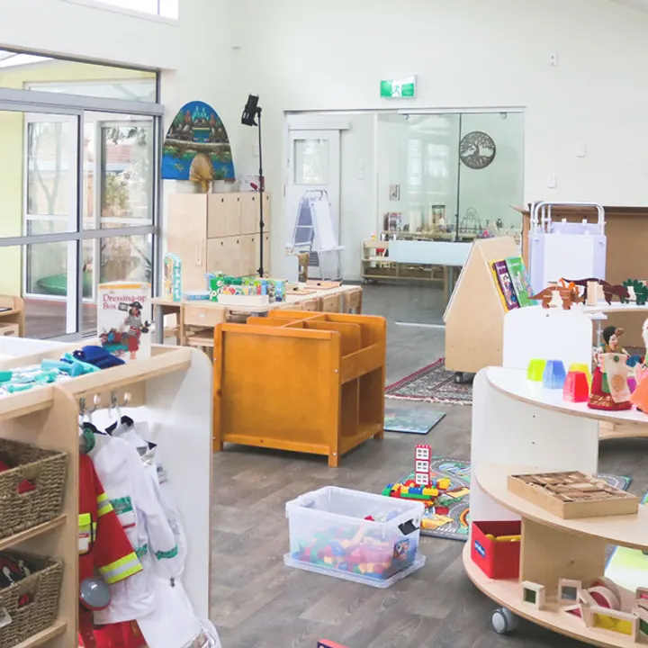 An open room set up with low wooden shelves, baskets of natural materials, a wooden trolley of blocks and a soft armchair.