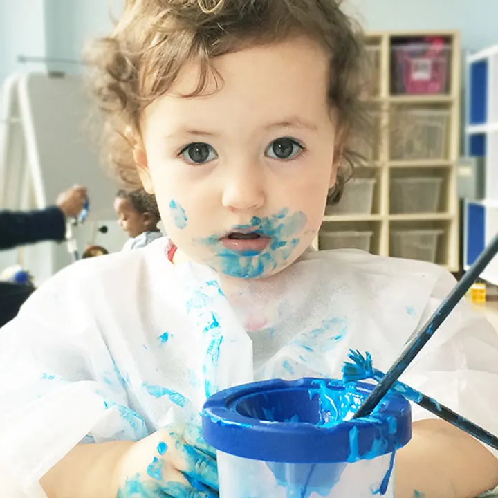 A toddler in a painting smock, hands and face covered in blue paint, holding a brush over a pot of it and looking up.