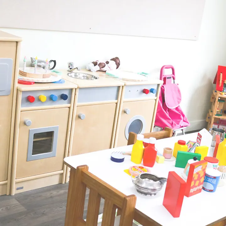 A wooden play kitchen with a sink, oven and washing machine, and a small table set out with plastic food and pots.