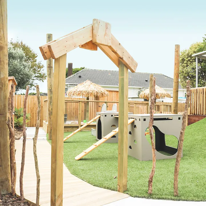 An outdoor play area with artificial grass, a timber archway, young trees and a climbing frame with a ramp and slide.