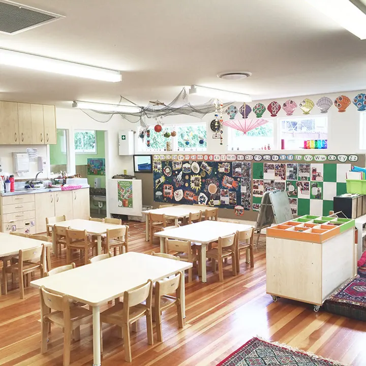 A large, bright preschool room with child-height wooden tables and chairs, a wooden floor, and children’s artwork covering the wall.